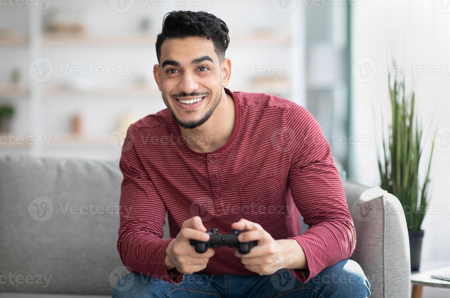 Emotional middle-eastern guy playing game with joystick at home, copy space. Happy millennial arab man enjoying weekend, sitting on couch in living room with joystick, gaming photo