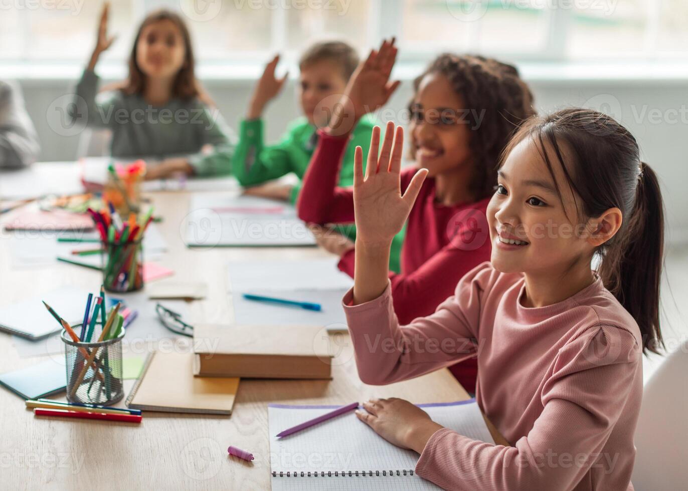 Happy Multiethnic School Kids Learning Raising Arms During Lesson Sitting At Desk In Classroom At School. Modern Education And Knowledge Concept. Selective Focus photo