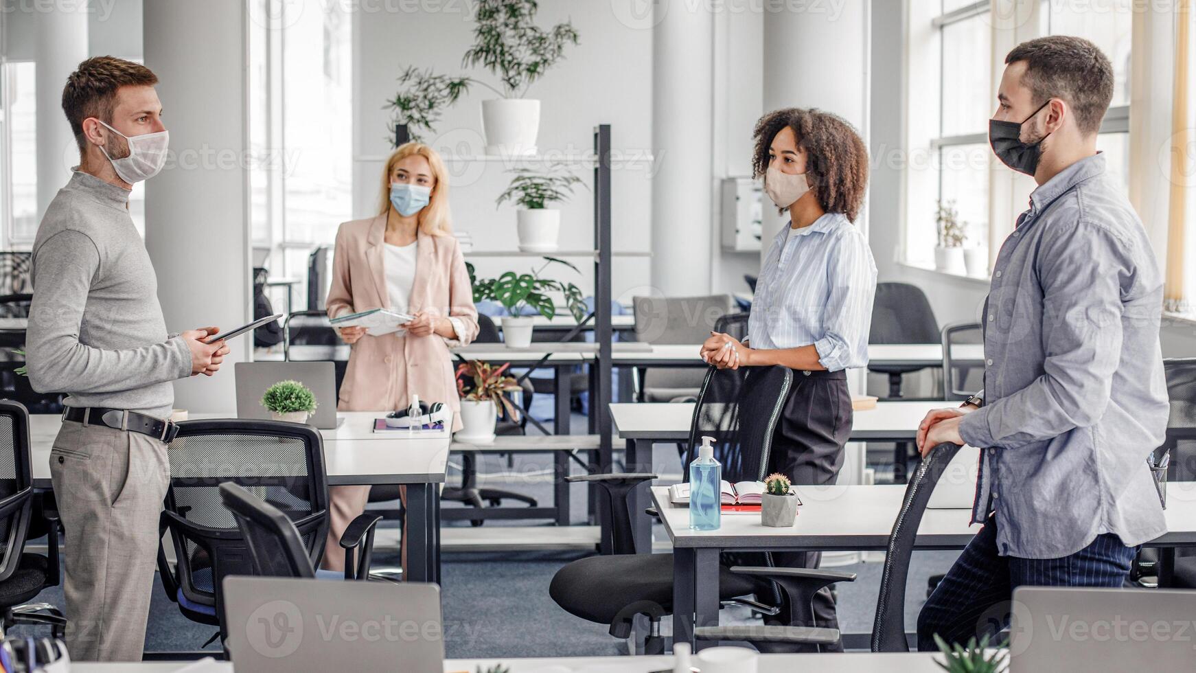Urgent meeting of colleagues about creative ideas and business plans. Multiracial colleagues in protective masks maintain social distance and discuss new normal at covid-19 in office interior photo