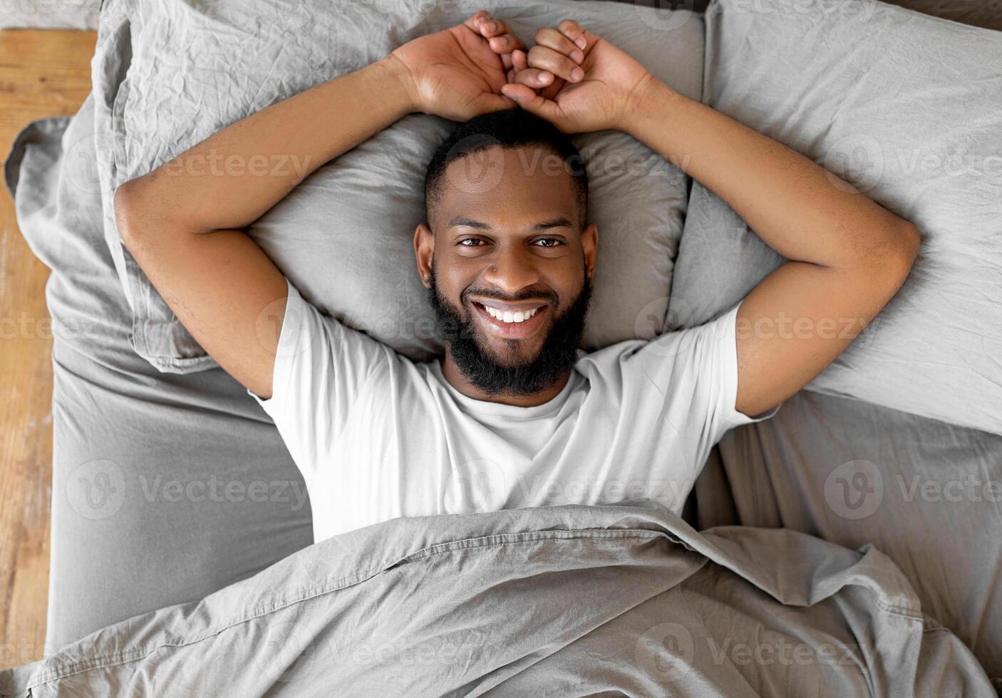 Overhead above top view portrait of relaxed smiling African American man lying on gray bedsheets at home after waking up, feeling good, peaceful young black guy resting in bedroom looking at camera photo