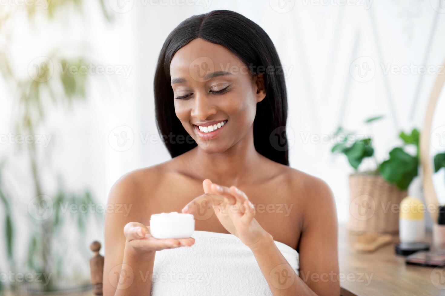 Home spa and permanent facial during covid-19 lockdown. Cheerful beautiful millennial african american female applying face cream to moisturize skin in bedroom interior in morning, copy space photo