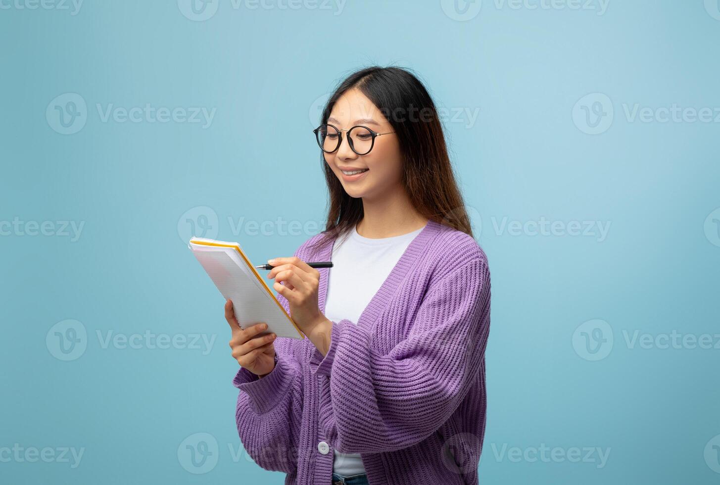 Intelligent asian lady in glasses taking notes, holding notepad and pen, standing over blue studio background, free space. Happy female student planning her day, writing tasks photo