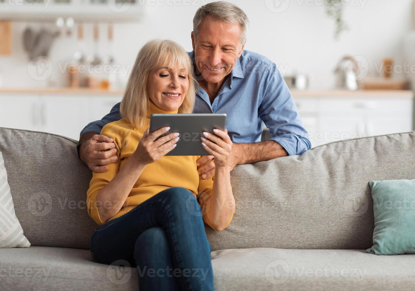 Happy Senior Couple Using Digital Tablet Together Browsing Internet Sitting On Sofa At Home. Retired Husband And Wife Reading Online News On Computer On Weekend. Mature People And Gadgets photo