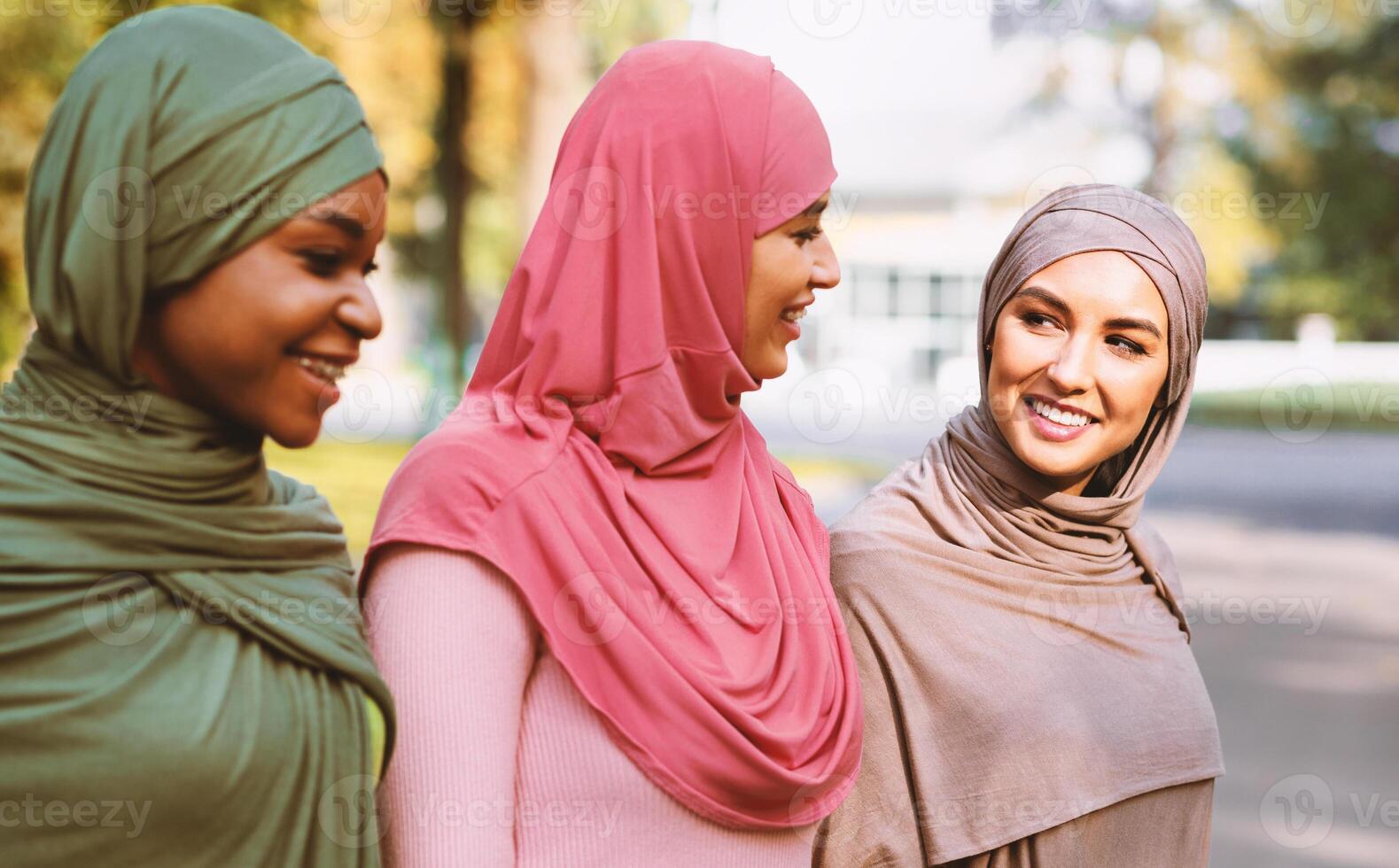Three Happy Muslim Women Walking Together And Talking In Park Outside Wearing Hijab Headscarf. Modern Islamic Females Lifestyle And Weekend Leisure Concept. Selective Focus photo