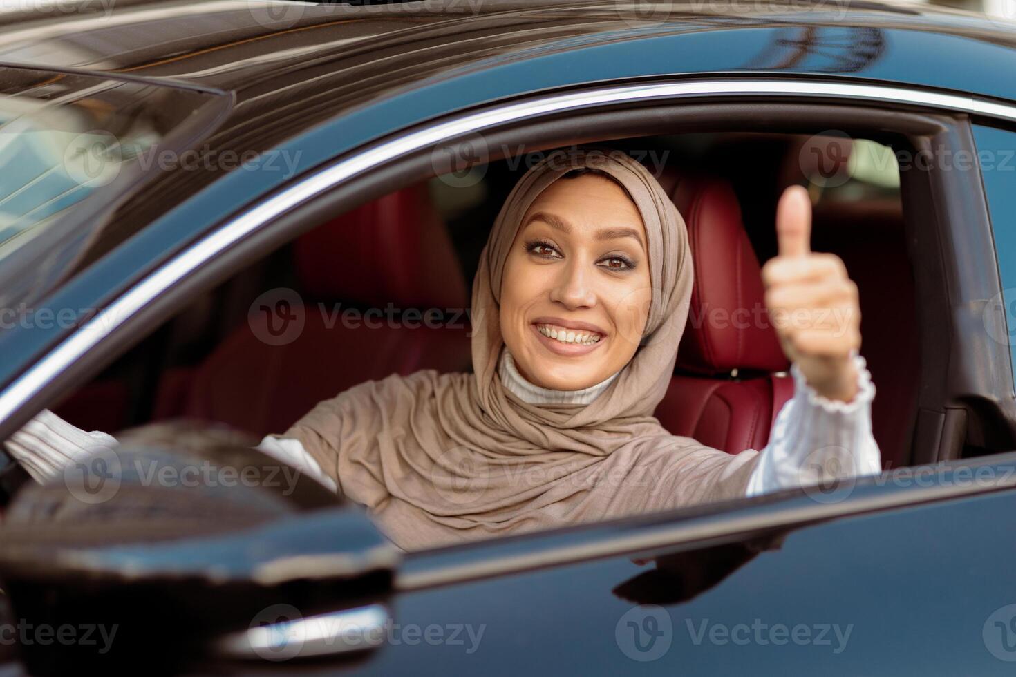 Great Car, I Like It. Portrait Of Smiling Muslim Woman In Hijab Gesturing Thumbs Up Sitting In Driver's Seat After Test Drive. Happy excited Arabian lady got driving license, gesturing okay sign photo