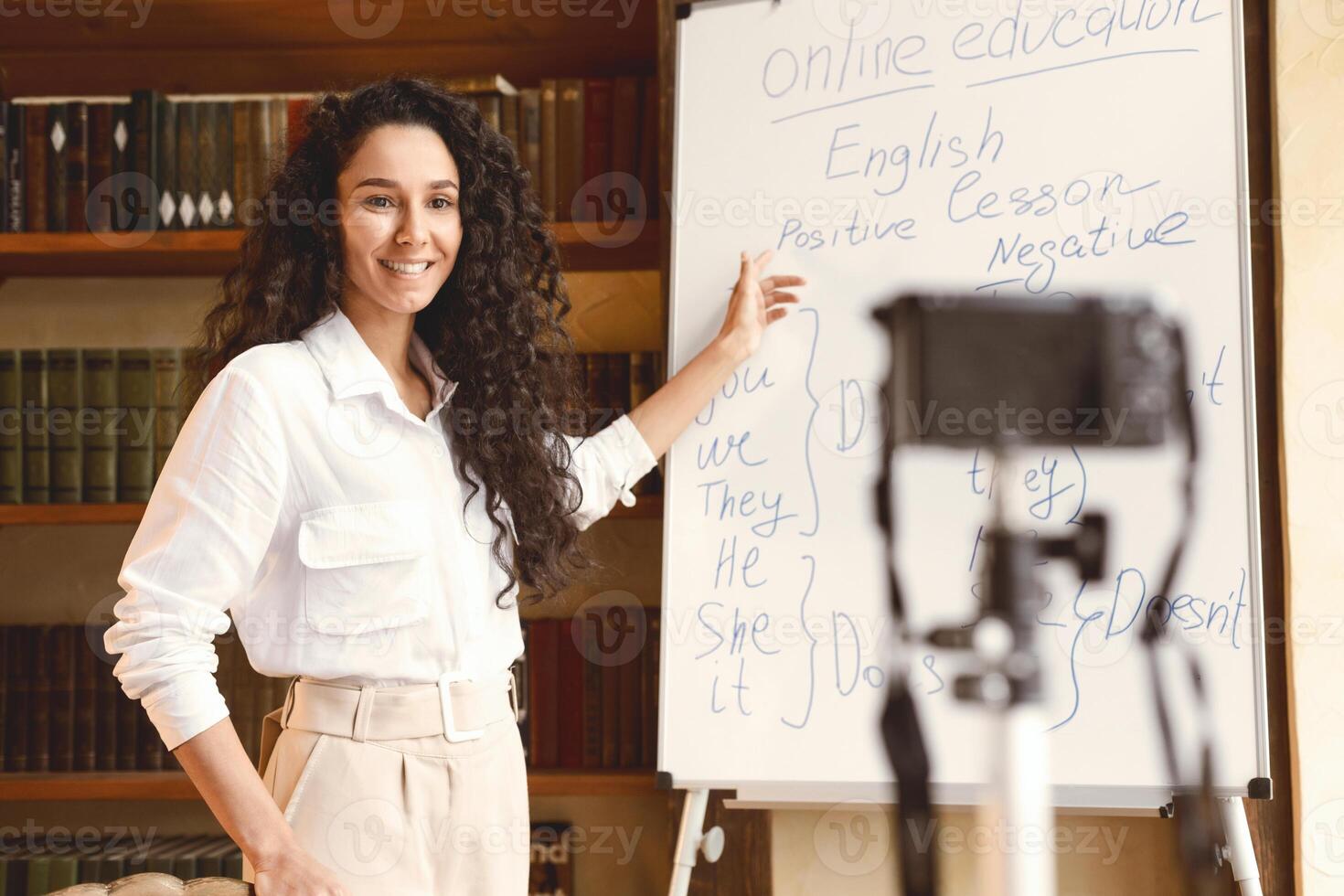 Remote Lecture. Young smiling female teacher explaining and giving online lesson, standing at whiteboard, filming her vlog. Woman recording blog for her internet channel at camera. E-learning photo
