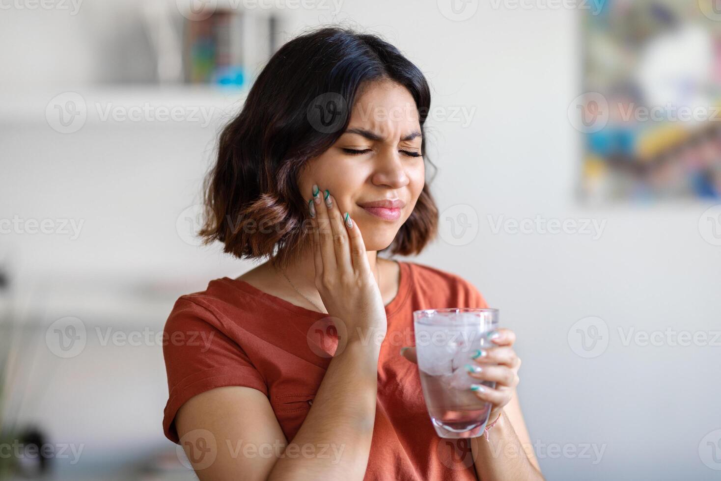 Sensitive Teeth. Young arab woman drinking water with ice and touching her cheek, sick middle eastern female frowning and rubbing inflamed area, suffering dental pain, emotionally reacting to cold photo