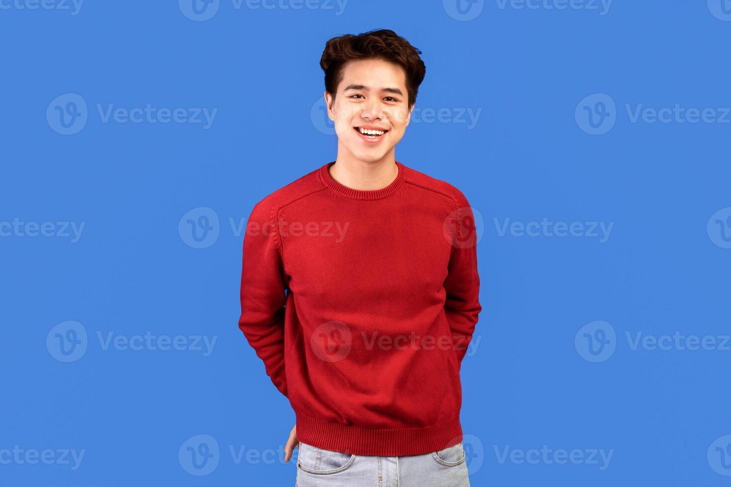 Male Portrait. Smiling asian guy standing and posing at studio, holding hands in pockets, looking at camera, wearing red sweater, posing isolated over blue studio background. Casual teenage model photo