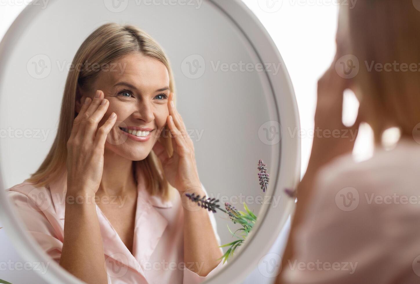 Beauty Concept. Portrait Of Attractive Mature Woman Looking At Mirror In Bathroom, Beautiful Middle Aged Lady Touching Her Flawless Face Skin And Smiling To Reflection, Selective Focus photo