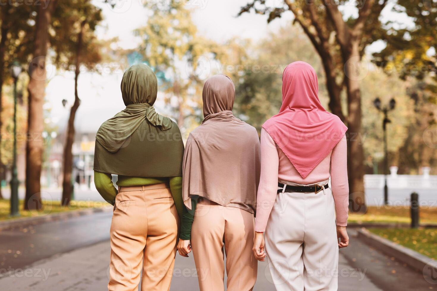 Back View Of Three Modern Muslim Women Walking Together Wearing Hijab Headscarves In City Park Outdoors. Islamic Females Friendship And Arabic Fashion Concept photo