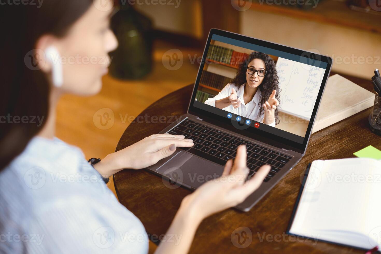Online Course And Distant Education Concept. Over the shoulder back view of young woman in earbuds sitting at desk, having online call with English teacher, talking and gesturing, using pc photo