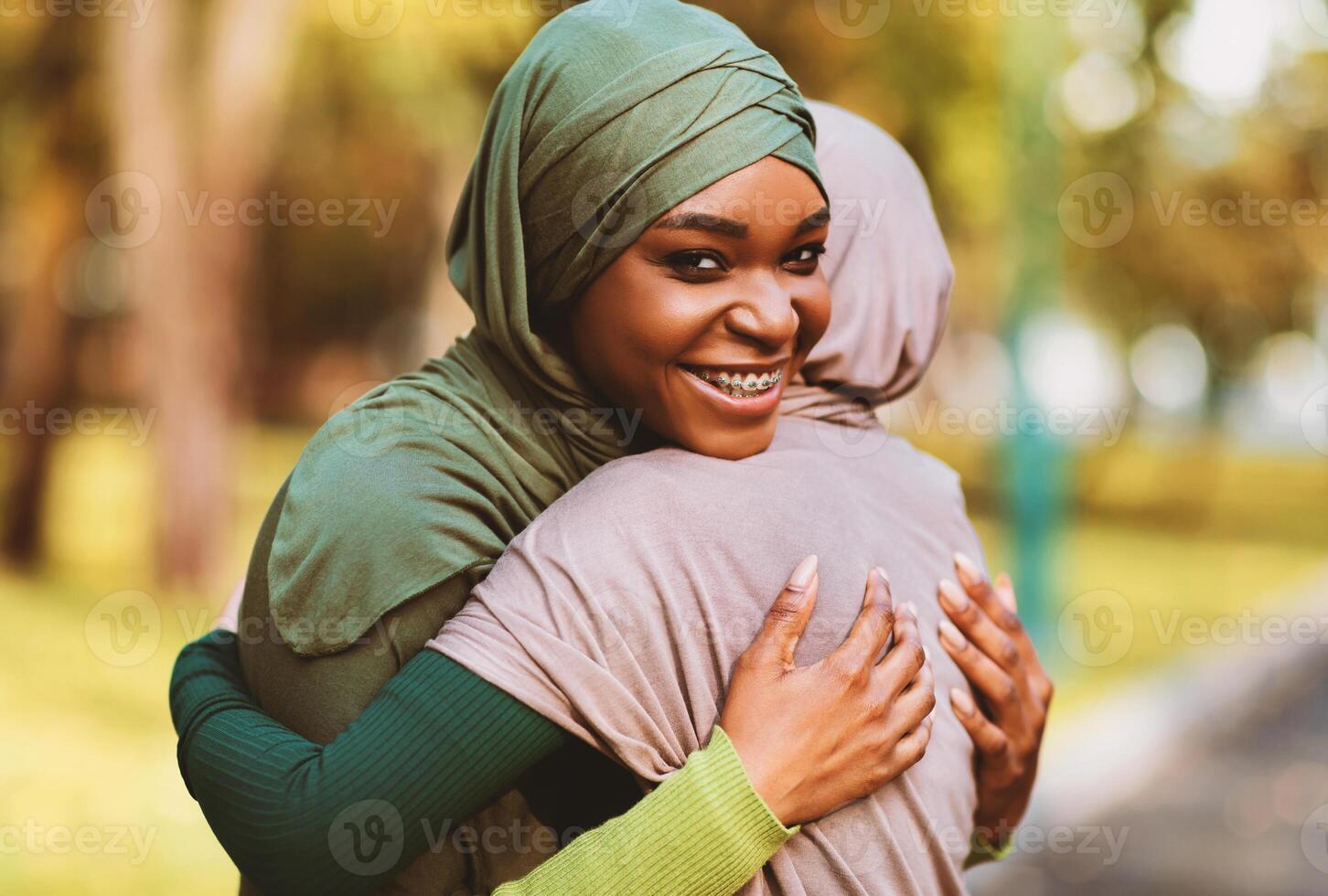Two Muslim Females Friends Hugging Smiling To Camera Standing In Park Outdoors. Cheerful Islamic Ladies Embracing Posing Outside. Unity, Friendship And Togetherness Concept. photo