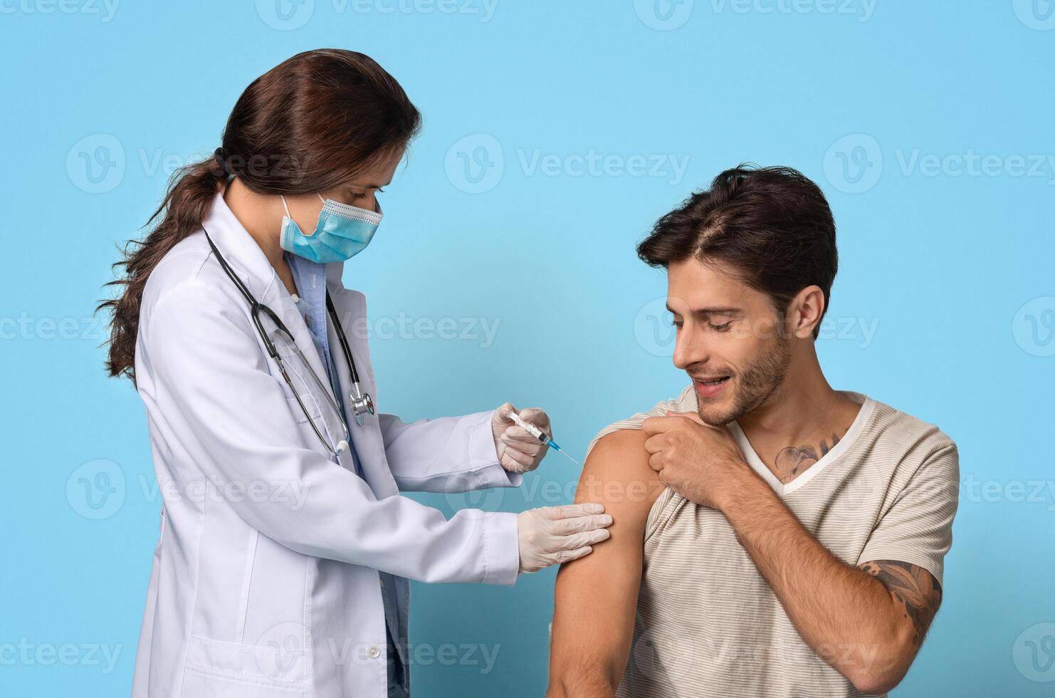 Vaccination Concept. Female Doctor With Syringe Making Vaccine Injection To Male Patient's Arm, Young Smiling Man Getting Vaccinated Against Covid-19 While Sitting Over Blue Background, Free Space photo