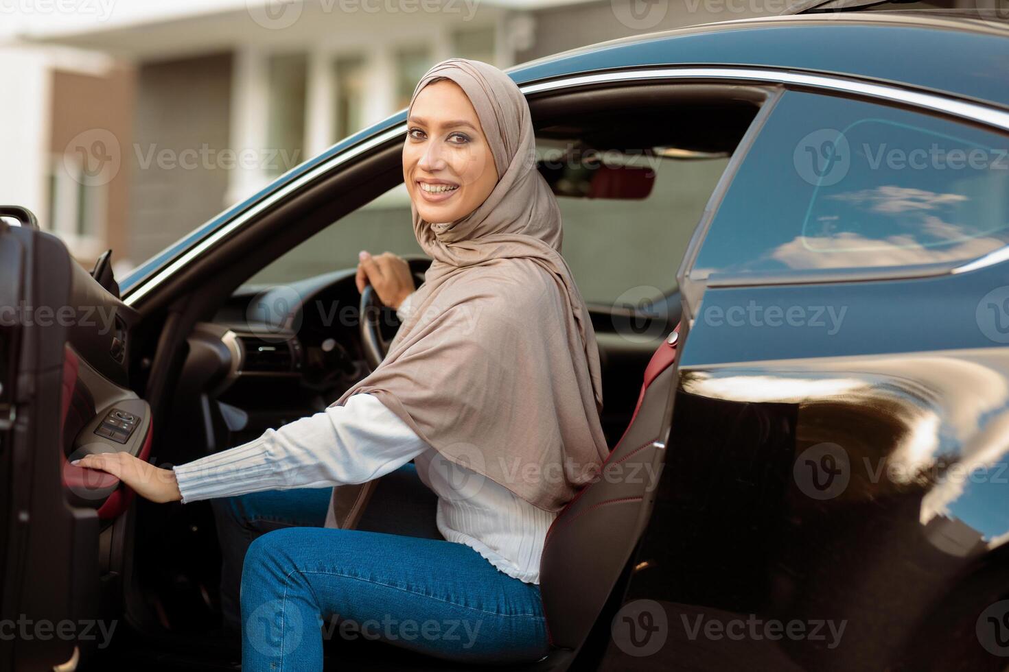 City Ride Concept. Portrait Of Happy Smiling Muslim Lady In Hijab Getting In Or Out Of Car, Holding Open Door. Arabian Woman In Headscarf Driving Modern Vehicle, Looking Back And Posing At Camera photo