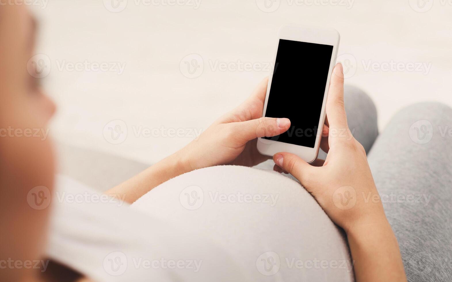 Unrecognizable pregnant woman using smartphone with blank screen sitting on sofa. Young expectant lady surfing net, reading about baby care, buying goods for newborn, copy space photo