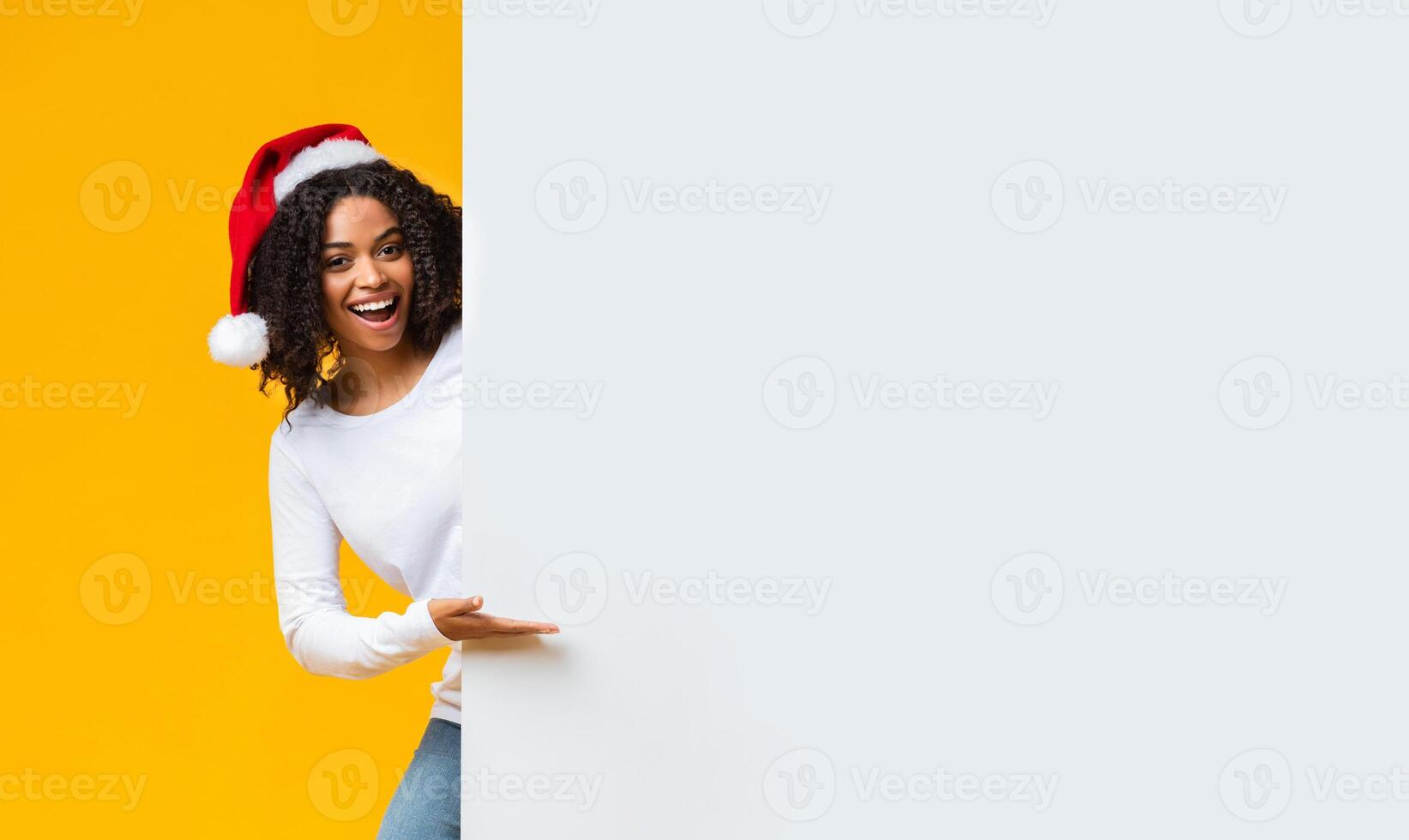 Christmas Offer. Happy African American Girl In Santa Hat Pointing At White Advertisement Board With Hand Over Yellow Background, Free Space photo
