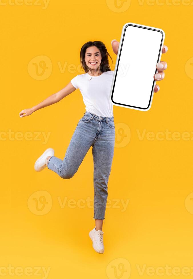 Beautiful young woman recommending new mobile application, vertical collage. Excited lady holding smartphone, showing white blank screen, jumping up over orange yellow studio background, full length photo