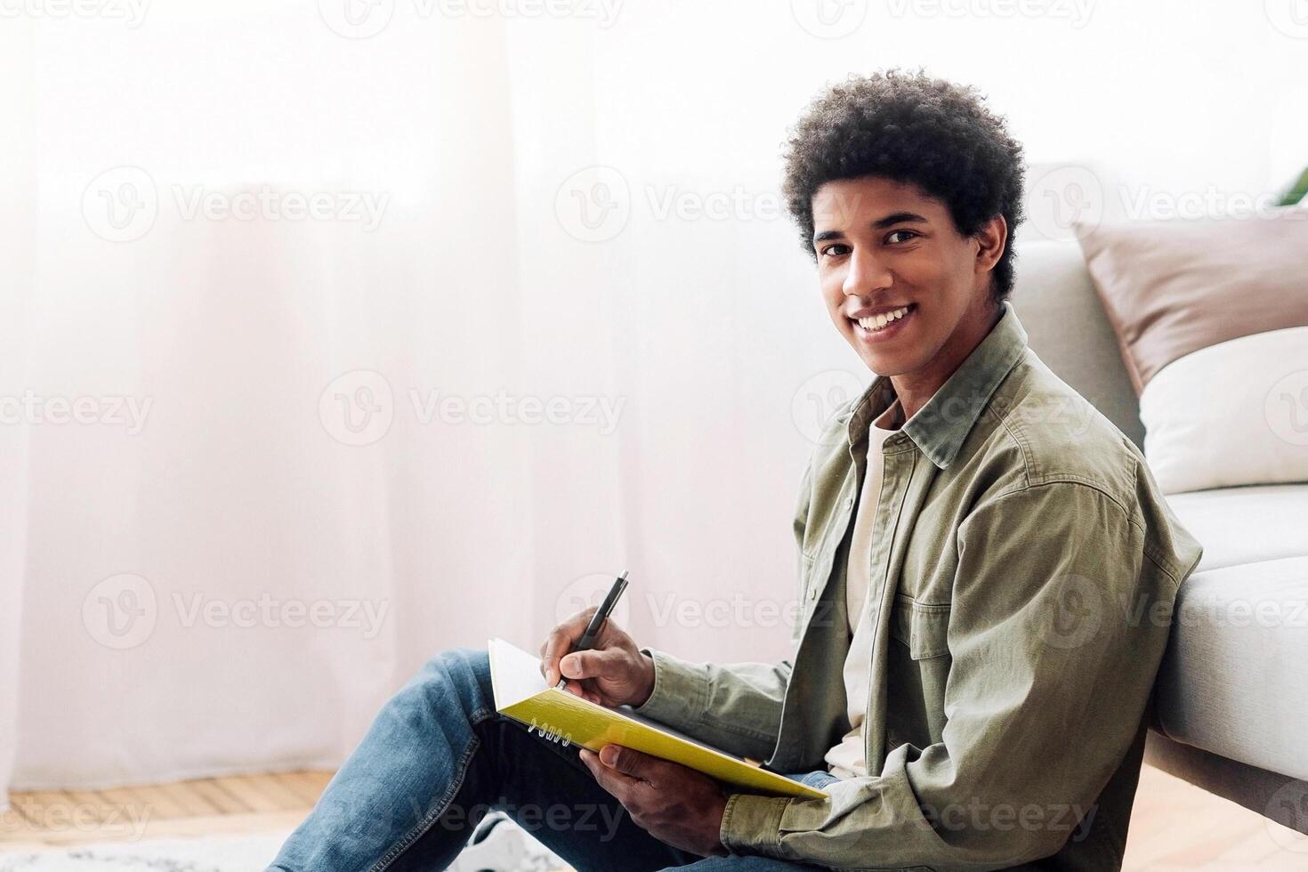 Black teen student with laptop taking notes for difficult exam during online studies at home, free space. Smiling teenage guy working on his assignment or coursework paper, making educational project photo