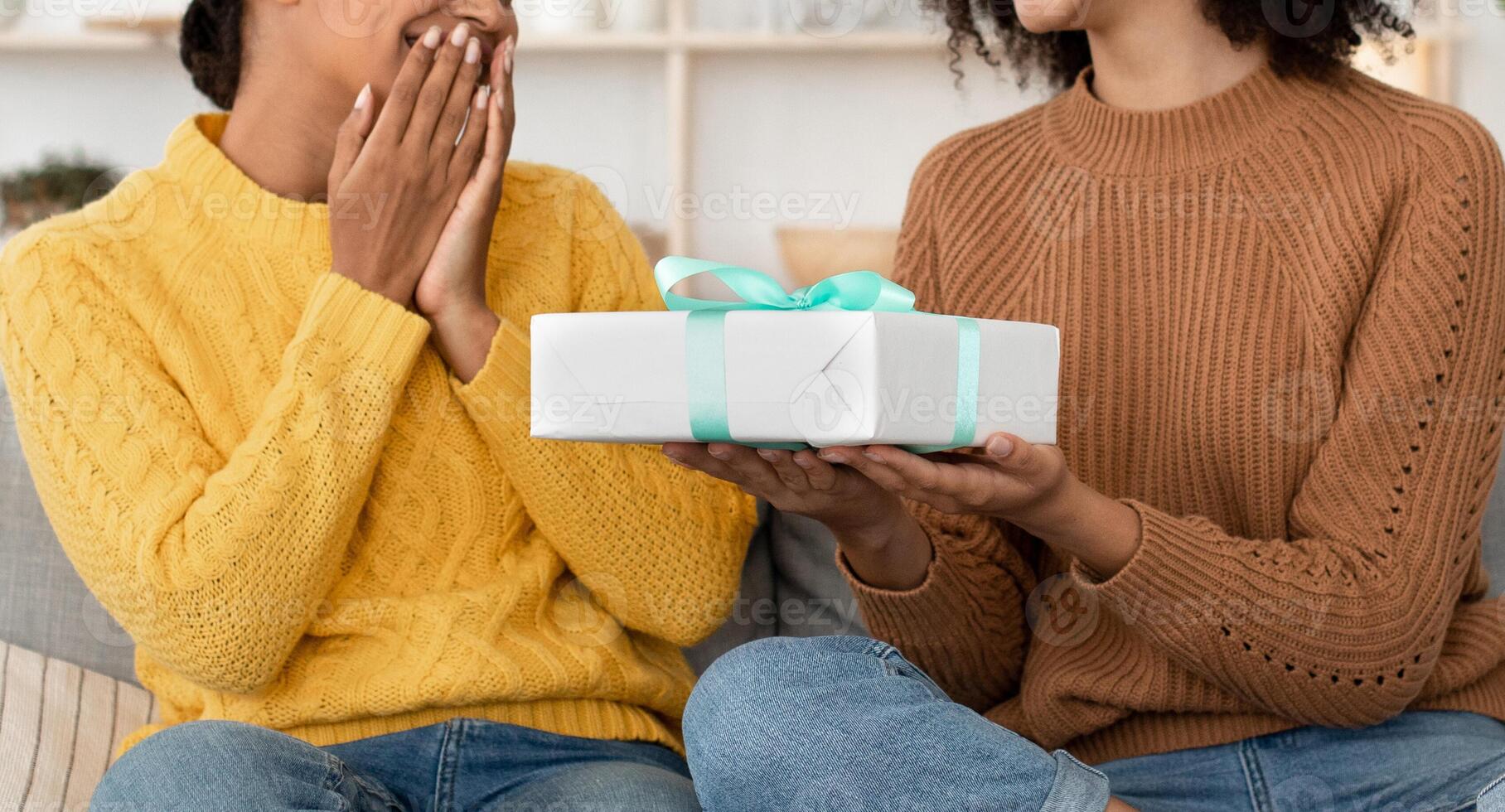 Gift for holiday, anniversary, birthday during covid-19 quarantine. Millennial african american female gives box with tape to surprised mixed race girlfriend, sits on sofa in living room, cropped photo