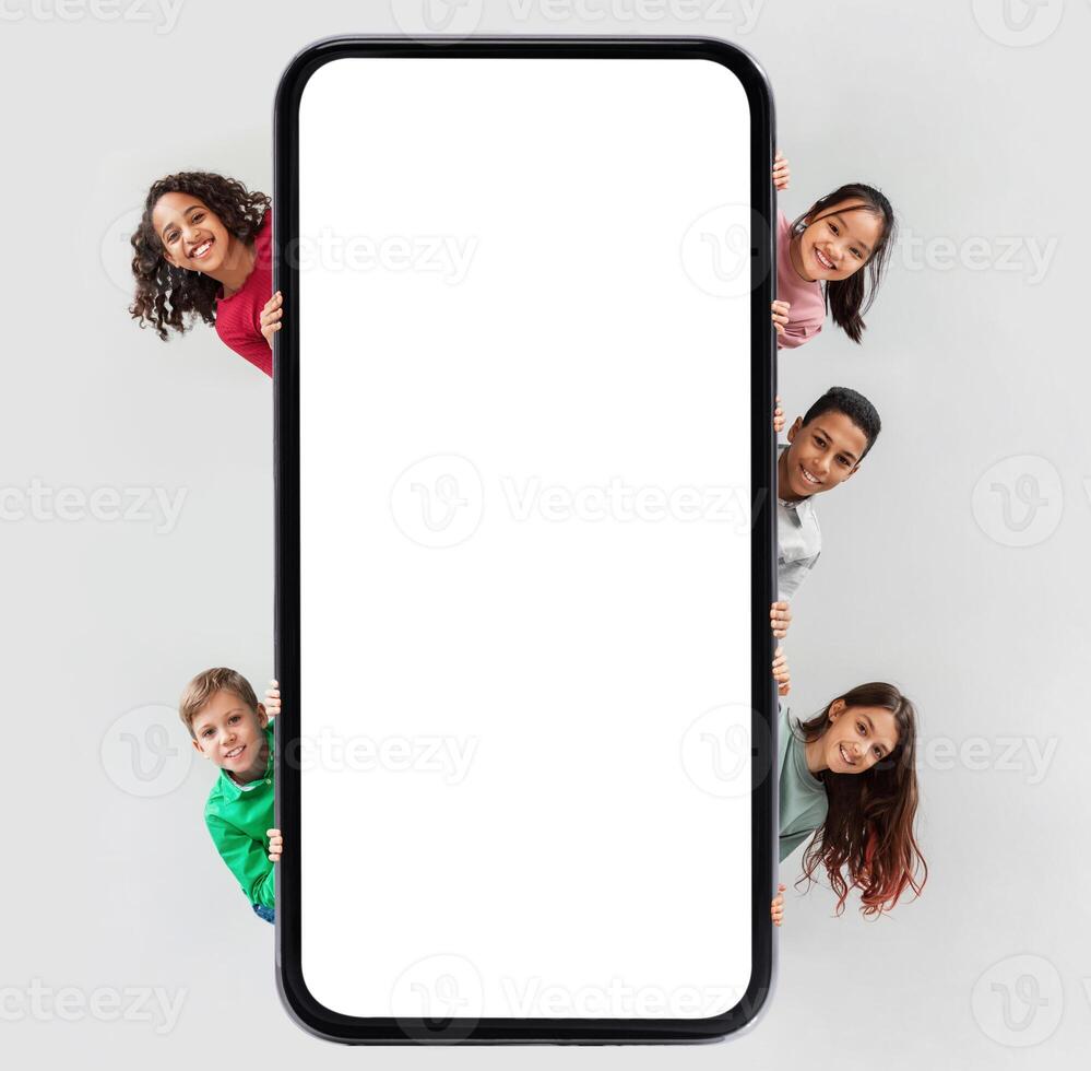 Diverse Kids Posing Near Huge Phone Blank Screen Hiding And Smiling To Camera Advertising Mobile Application Over Gray Studio Background. Children And Gadgets Concept. Square Shot, Mockup photo