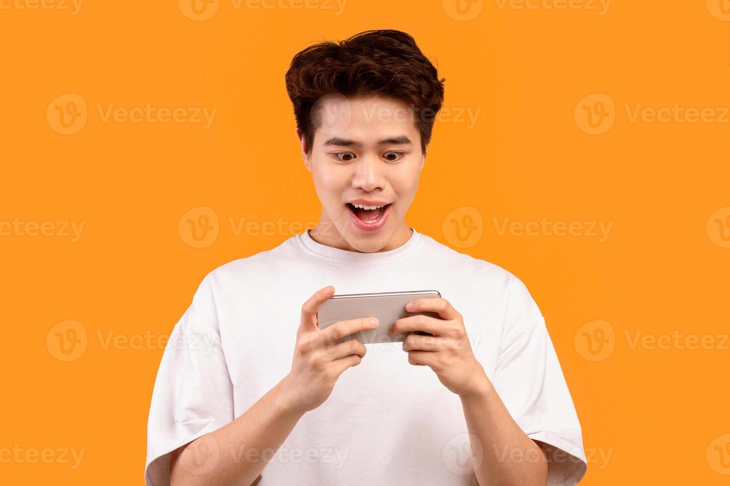 Great App. Portrait of excited joyful asian teen playing mobile games online on cellphone isolated over orange studio background, copy space. Emotional guy watching , browsing internet photo