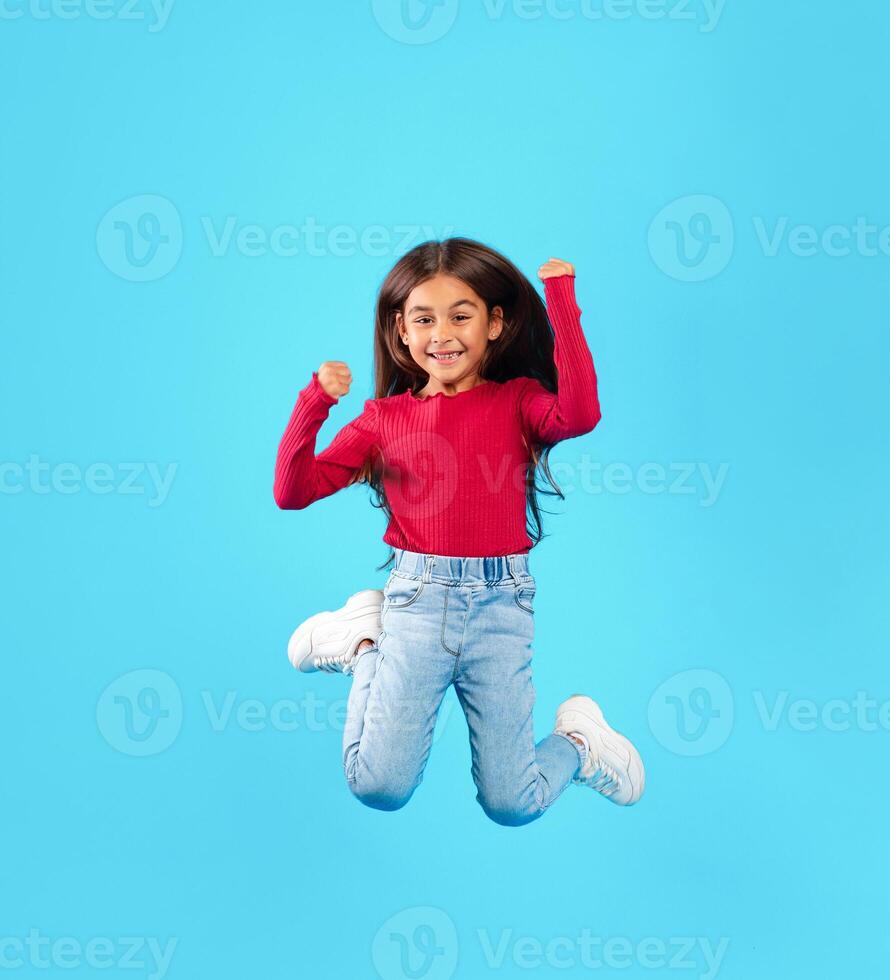 Happy Little Girl Jumping Having Fun Posing In Mid-Air Smiling To Camera Over Blue Studio Background. Full Length Shot Of Carefree Kid Celebrating Success. Vertical photo