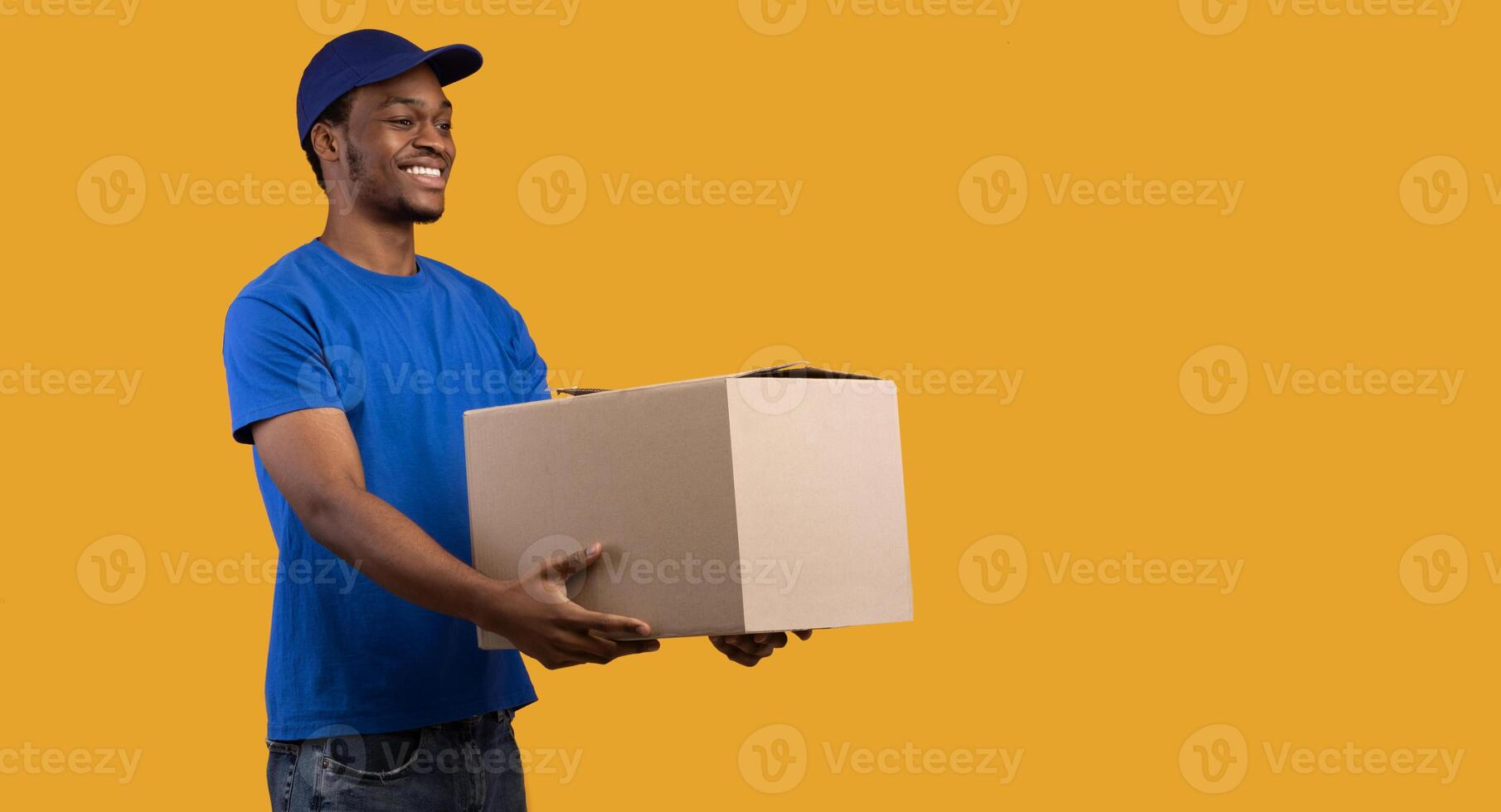 Portrait of cheerful african american delivery guy loader wearing blue cap and uniform holding cardboard container, giving it to the side, copy space, banner. Man isolated on orange studio background photo