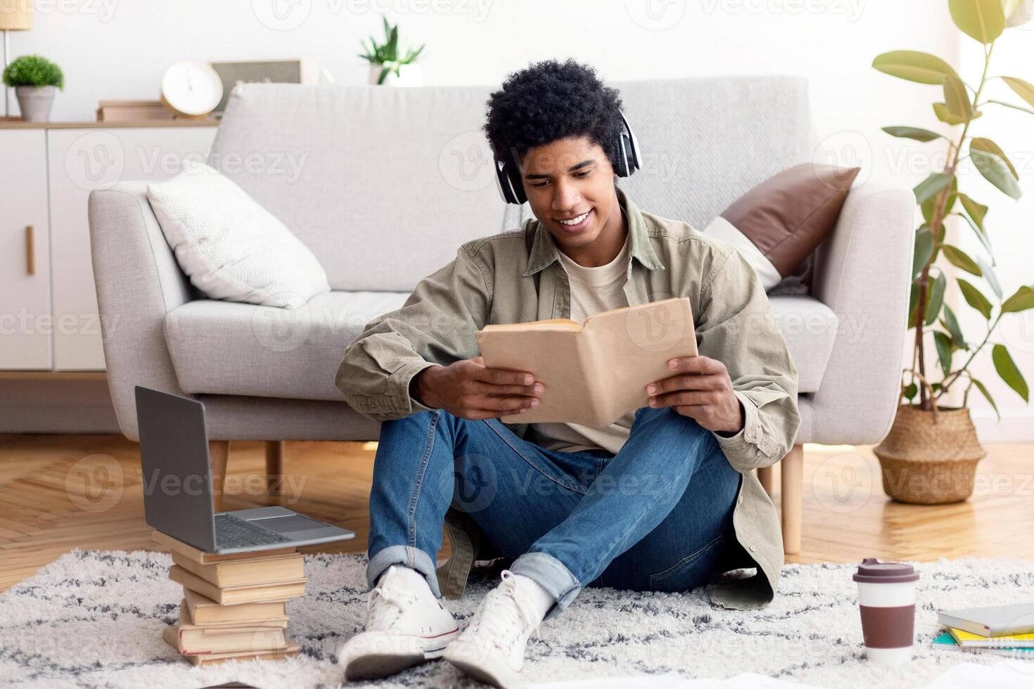 Busy teenage male student in headphones reading book during online class near laptop computer indoors. Positive black guy studying remotely, taking part in web conference, listening to lecture at home photo