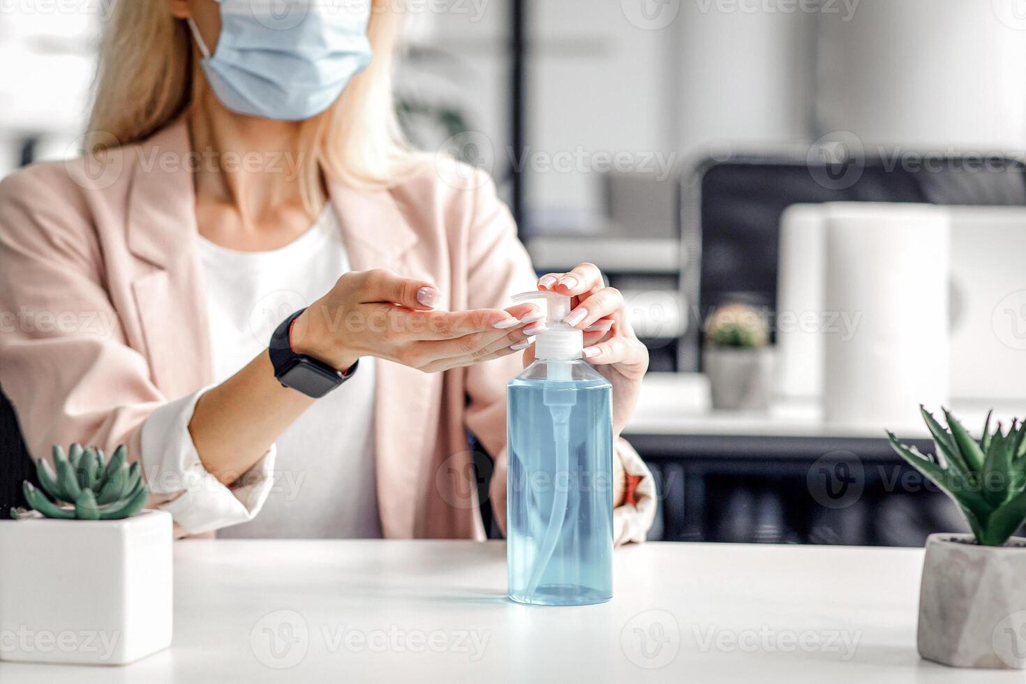 Safety and health protection at work in office. Millennial woman in protective mask with smart watch uses antiseptic at workplace with laptop for hand disinfection during covid-19 pandemic, empty space photo