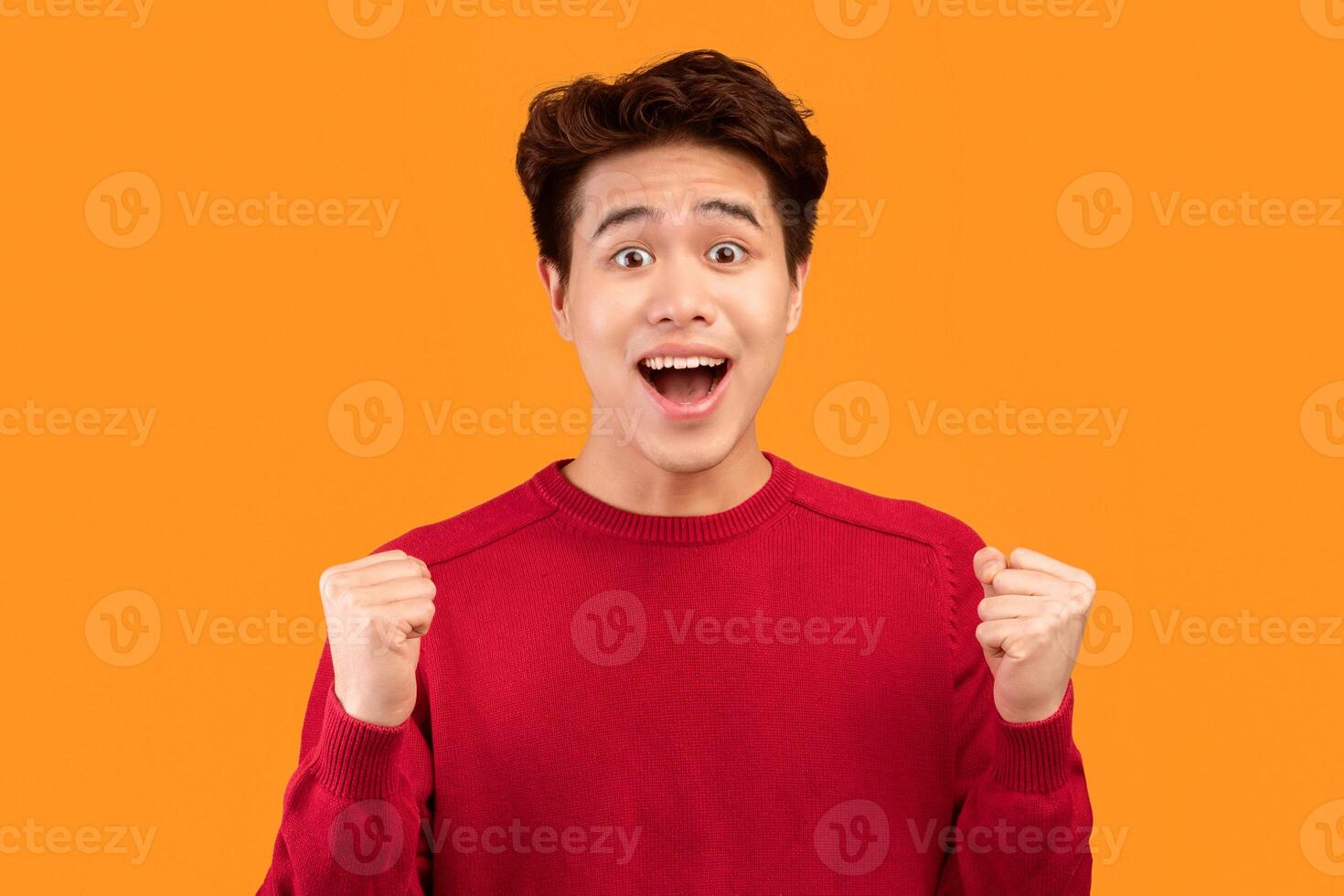 Omg, Great News. Portrait of joyful asian guy screaming and shaking clenched fists raised up in the air, isolated over orange studio background. Positive teen cheering and celebrating victory or win photo