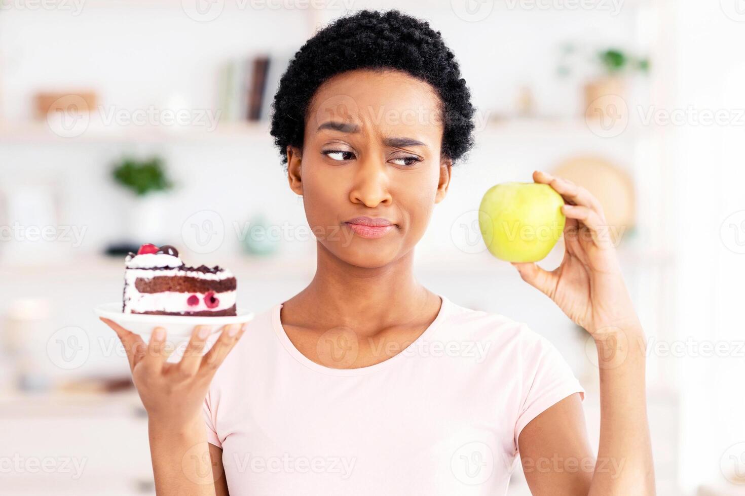 Choosing healthy or unhealthy food. Young black woman looking at apple with suspicion, not wanting to eat fruit instead of cake at home. Millennial lady preferring sweets, tired of weight loss diet photo