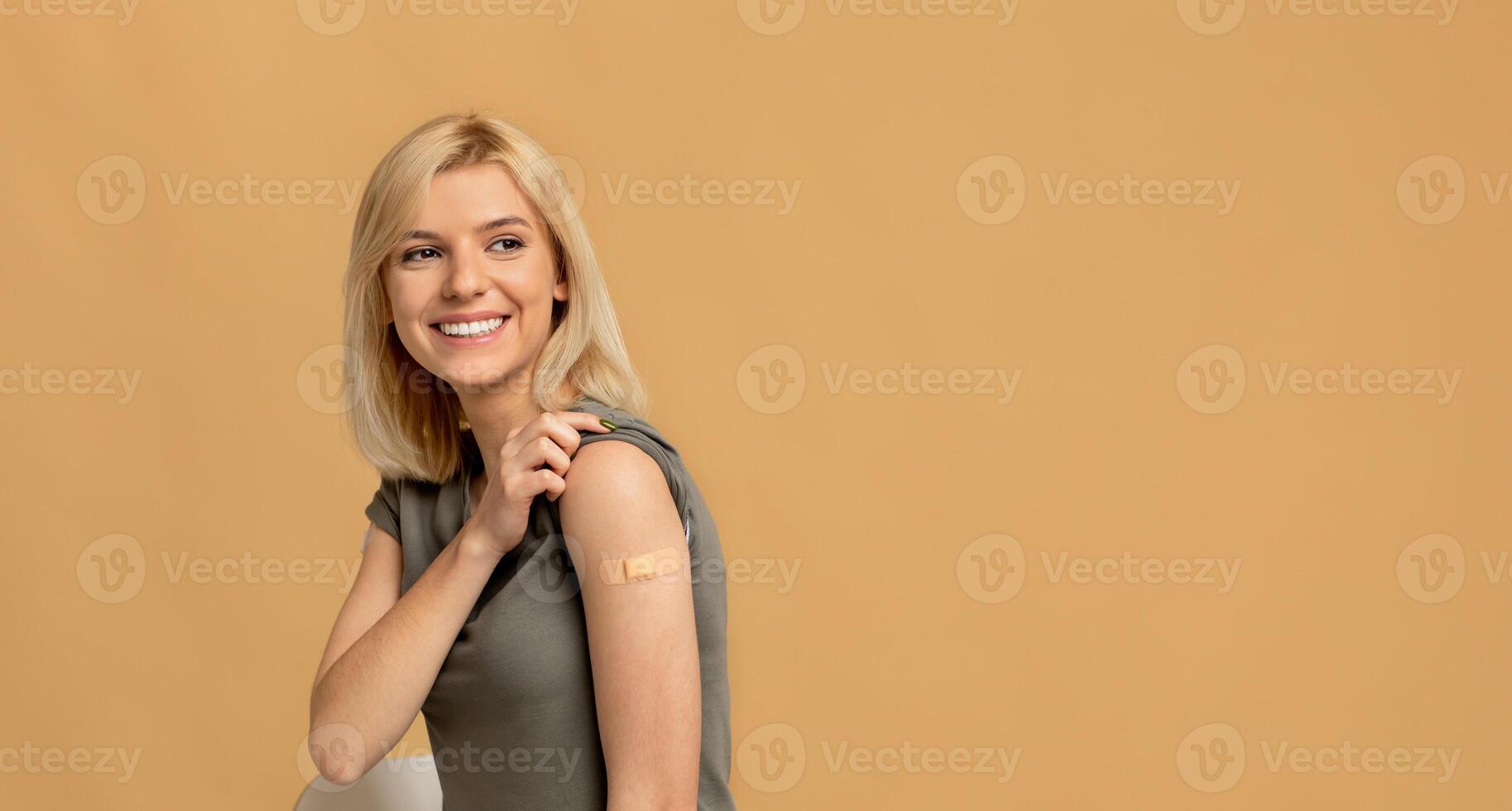 Vaccination saves lives. Happy woman getting vaccinated immunity to covid-19, posing over beige background with empty space, panorama. Female patient showing shoulder with band aid after shot photo