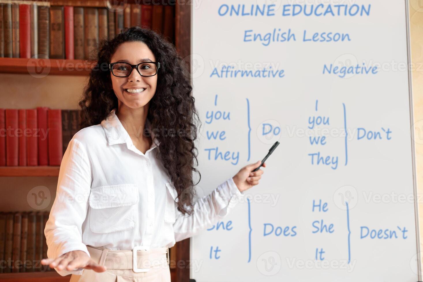 Online Education Concept. Smiling professional English teacher standing and pointing at whiteboard with marker. Young female lecturer wearing eyeglasses, explaining grammar rules to the students photo