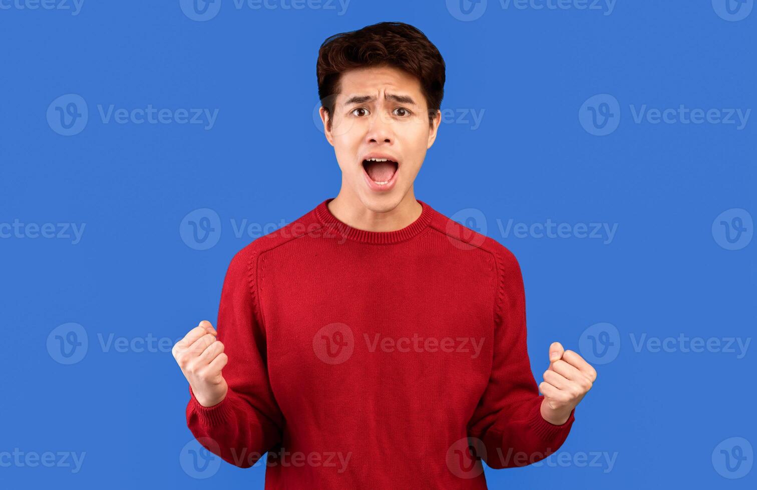 People, Human Emotions And Reaction Concept. Portrait of emotional asian young man screaming, shouting and shaking clenched fists raised up in the air, isolated over blue studio background photo
