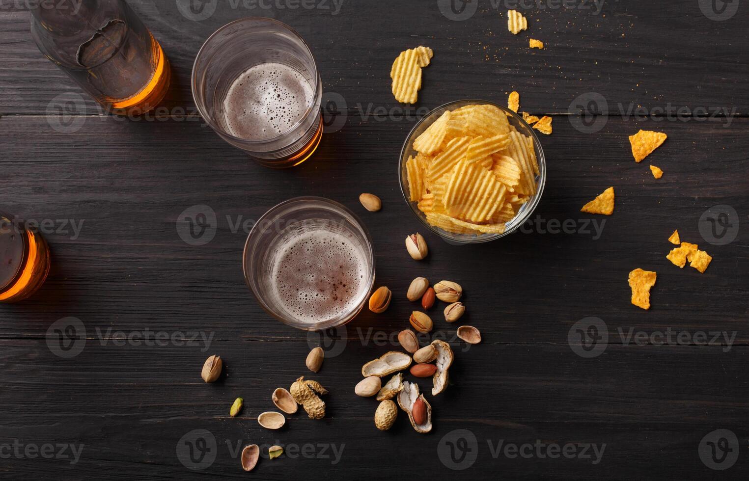 Beer event. Chips and pistachios in glass bowl on dark table, near are two glasses with light lager and bottles, top view photo