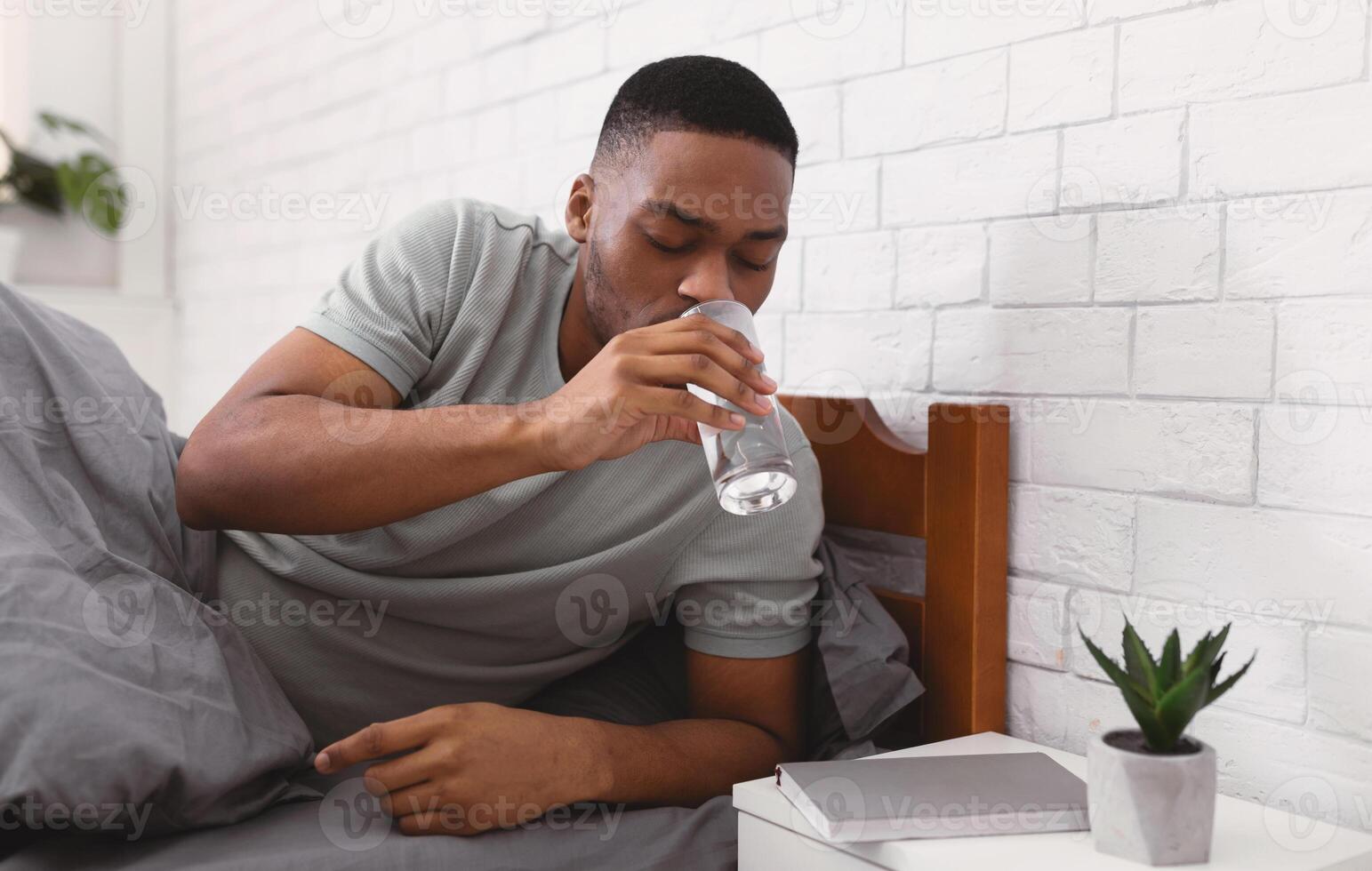 Black Man Taking Glass Of Water Lying In Bed, Hydrating Drinking In Bedroom At Home In The Morning, Starting New Day With Fresh Aqua. Stay Hydrated. Healthy Liquid Concept. Selective Focus photo