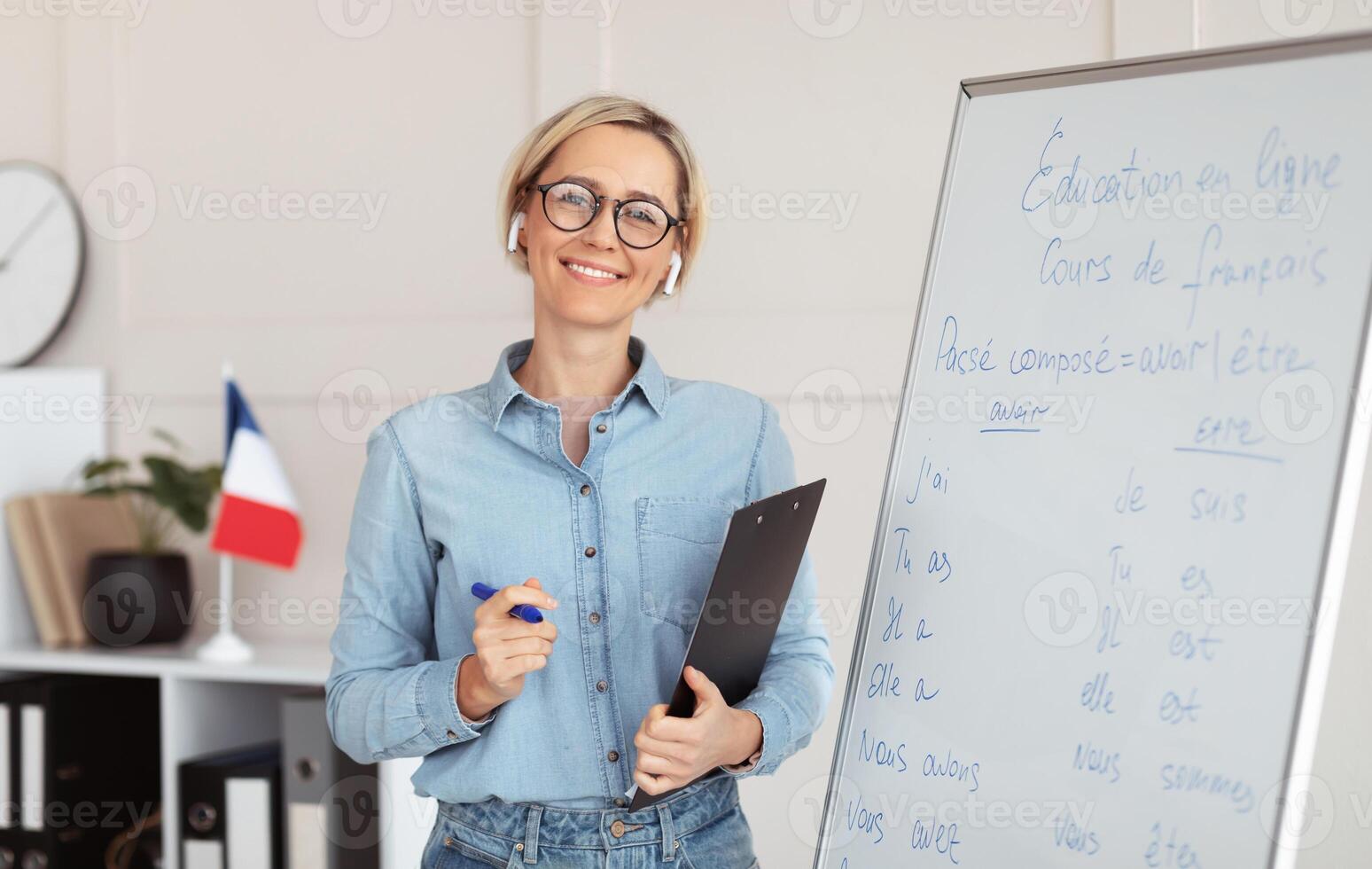 Friendly young female tutor standing near blackboard with French grammar rules, giving web lesson from home. Foreign language professor teaching students online. Remote education concept photo