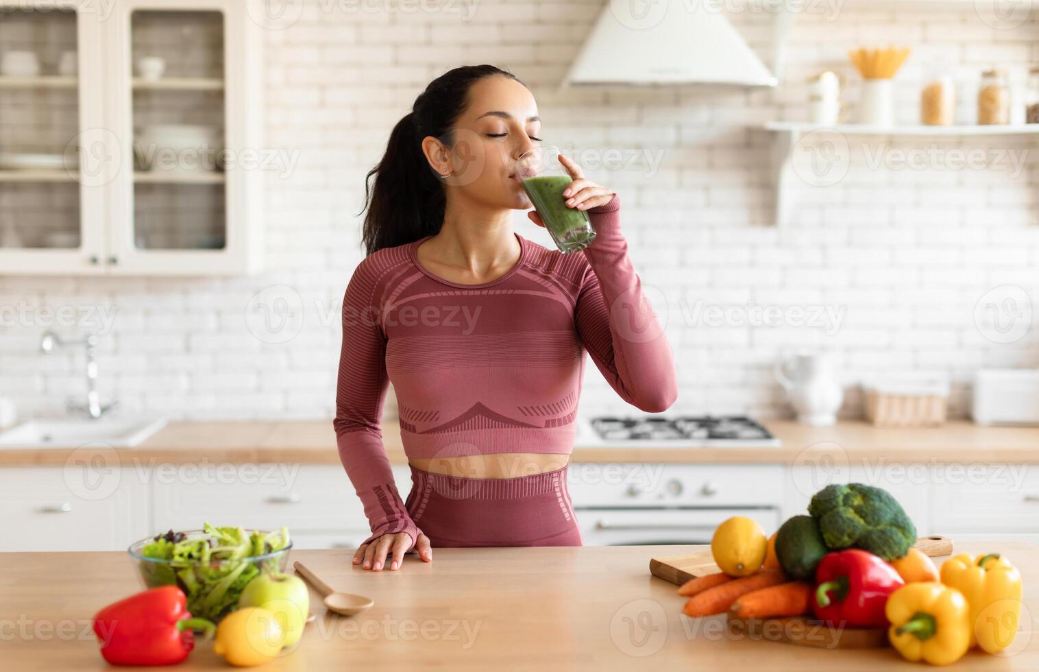 Sports Nutrition. Slim Young Woman In Fitness Fitwear Drinking Green Smoothie, Enjoying Detox Drink Posing With Glass At Modern Kitchen At Home, Standing Near Table With Fruits And Vegetables photo