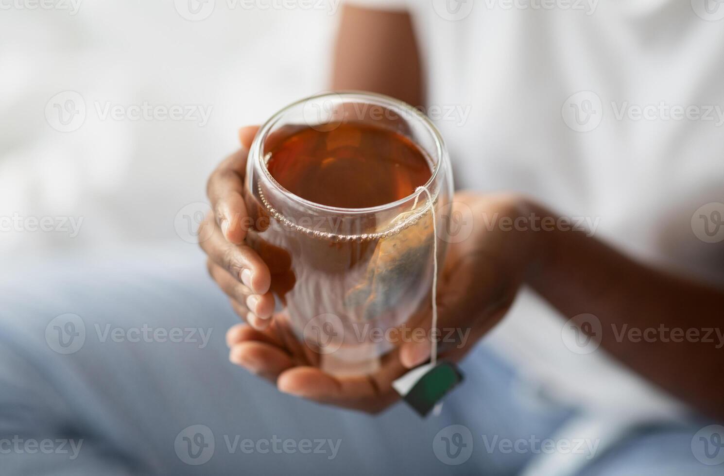 Unrecognizable black woman in pajams sitting on bed and holding glass mug with herbal or healthy green tea, enjoying refreshing hot drink in the morning after waking up, cropped, copy space photo