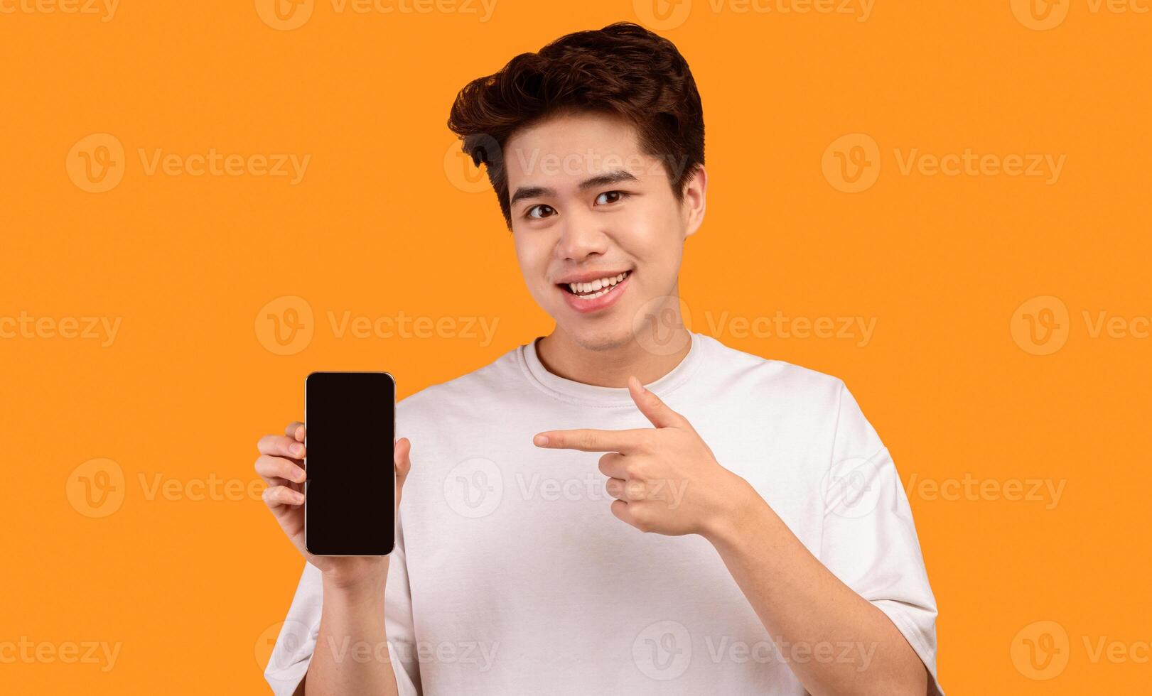 I Recommend This Website. Smiling young asian man holding smartphone with black empty screen in hand and pointing at device. Gadget with blank space for mockup, isolated over orange studio background photo