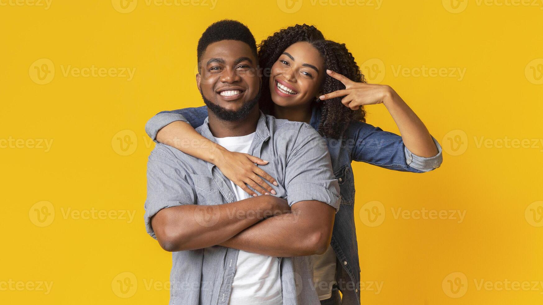 Portrait of millennial afro couple embracing and posing at camera, woman showing peace gesture, yellow studio background, panorama photo