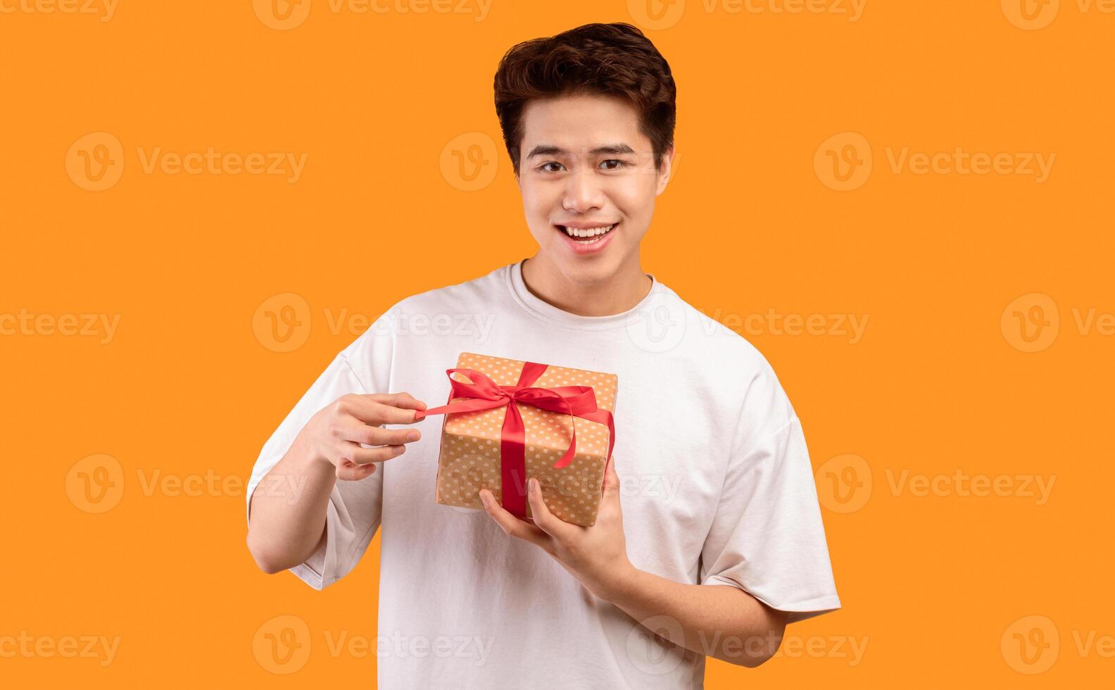 Holiday Concept. Portrait of smiling asian guy holding gift box, unwrapping and pulling red bow ribbon, looking at camera, isolated over orange studio background, greeting and celebrating special day photo