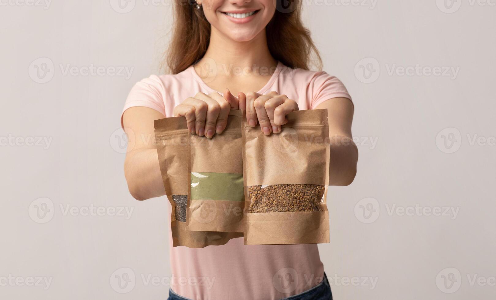 Eco-Friendly Package. Unrecognizable woman holding brown kraft paper doypack bags with weight food groceries in hands, female buyer standing on grey background in studio, cropped image, closeup photo
