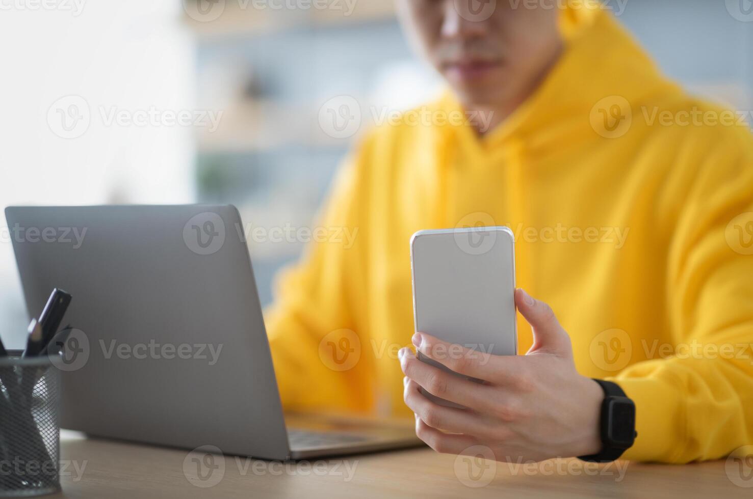 Closeup of unrecognizable man wearing yellow hoodie using phone and laptop, working on online project sitting at desk in modern home office, young employee looking at smartphone screen, reading email photo