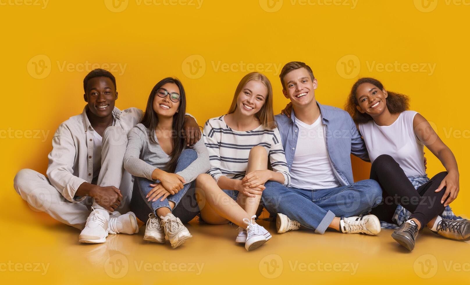Multiracial joyful students sitting on floor, embracing and having fun over yellow studio background, students lifestyle concept, panorama photo