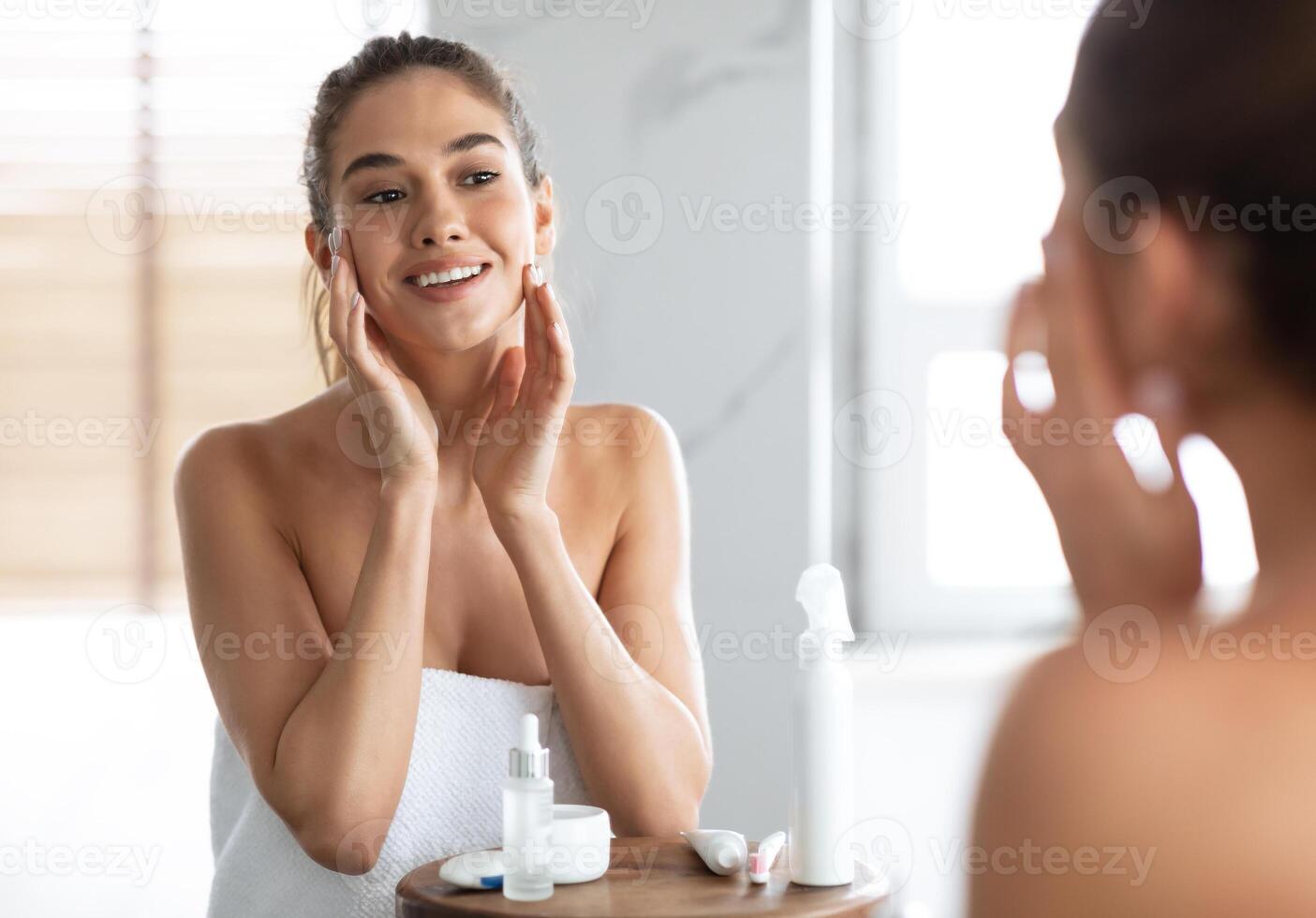Perfect Skin. Happy Woman Touching Face Caring For Young Skin Looking In Mirror Standing In Bathroom At Home, Wrapped In Towel. Female Skincare Beauty Routine Concept. Selective Focus photo