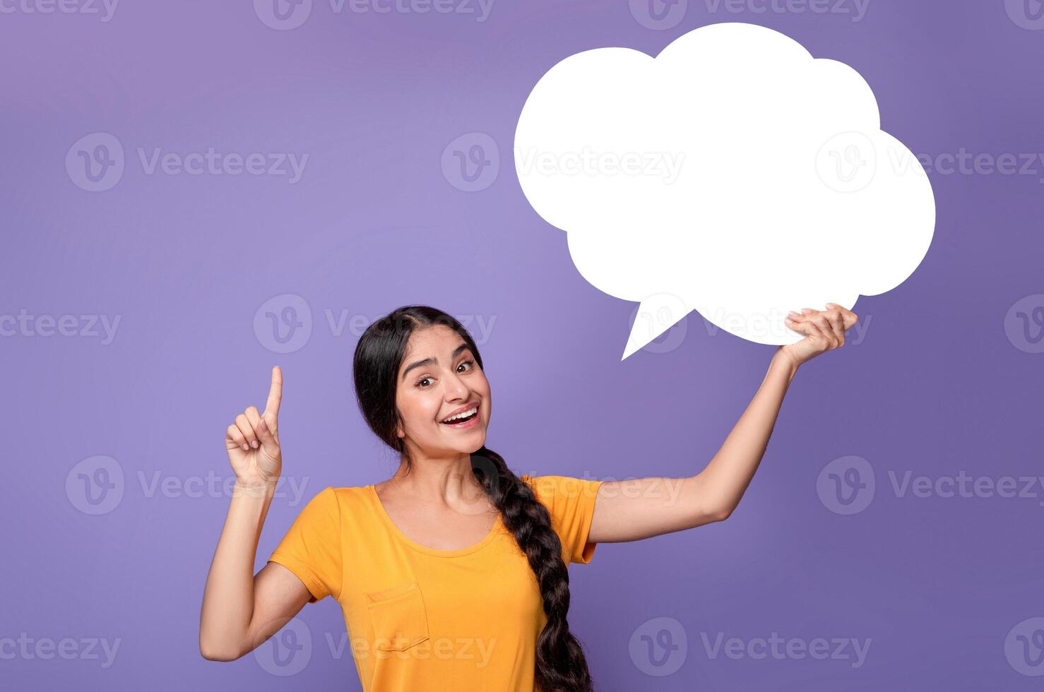 Inspired young indian lady having idea, holding blank speech bubble and pointing finger up, showing free place for your text, standing over purple background in studio, copy empty space photo