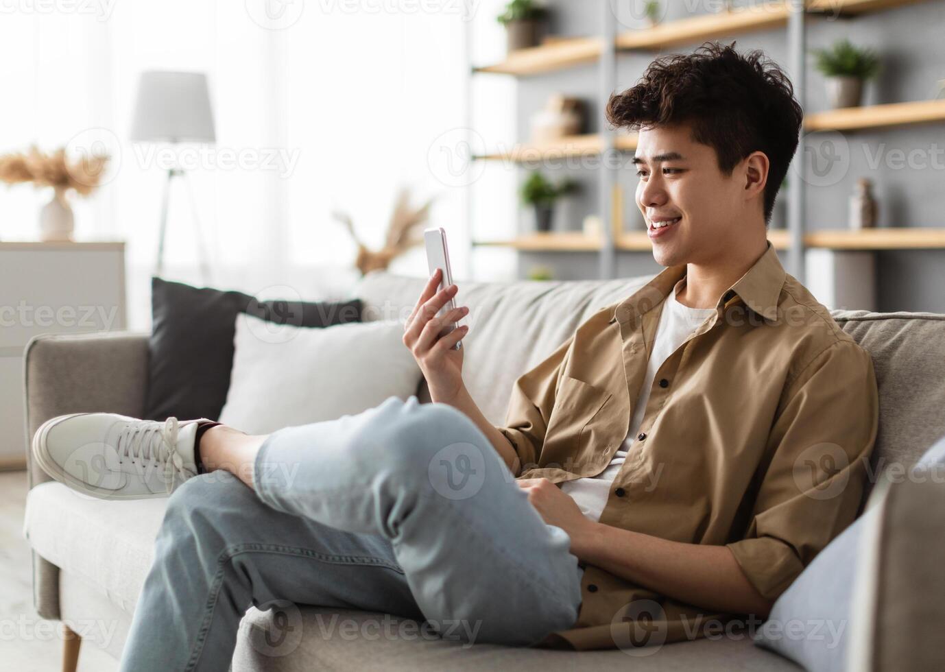 Cool Gadget And Application. Portrait of young smiling asian man holding mobile phone, typing sms message, sitting on the sofa in living room. Guy browsing internet, surfing web, using app, free space photo