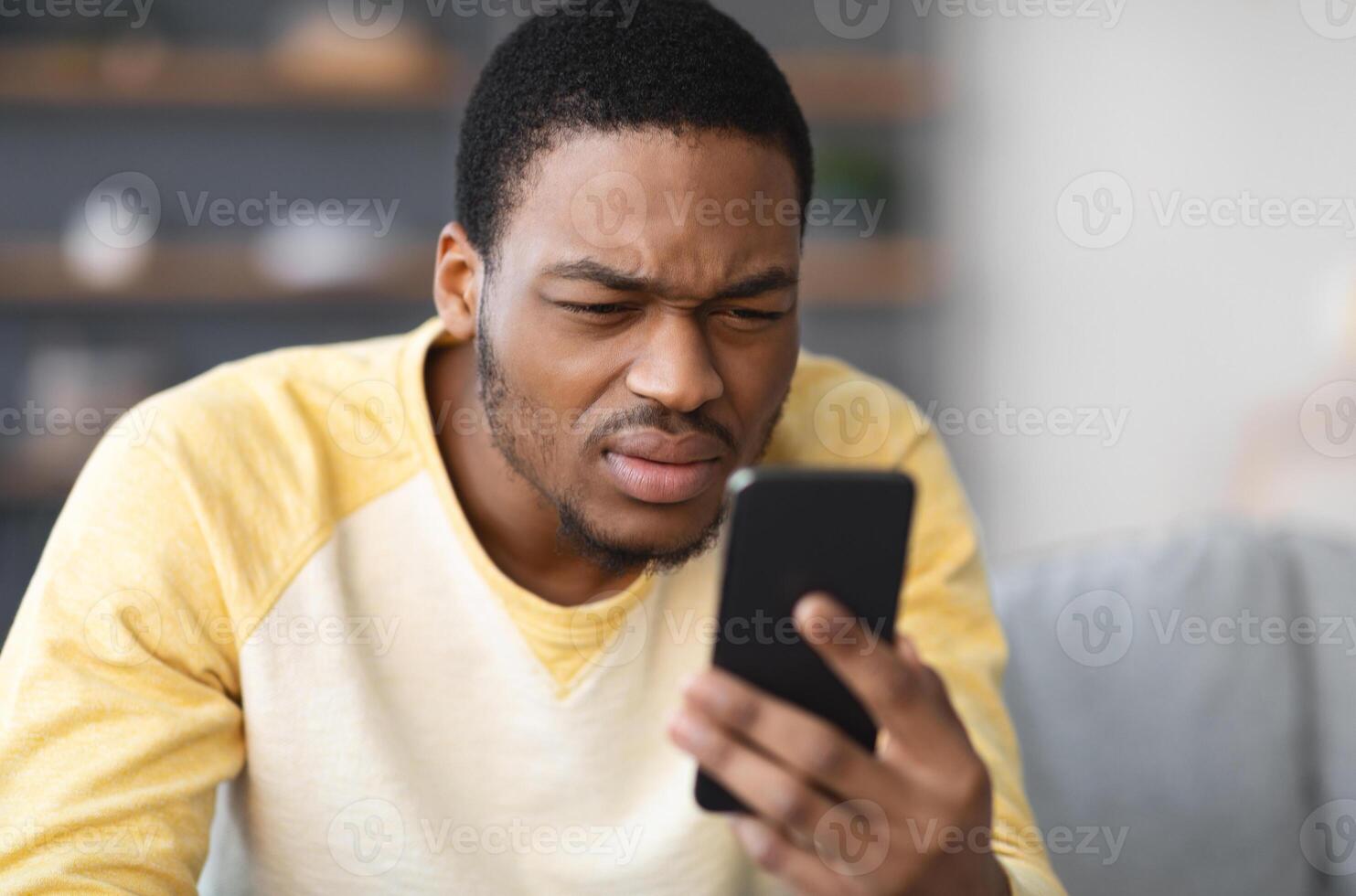African american guy squinting while looking at mobile phone screen at home, copy space, blurred background. Young black man reading suspicious message on his smartphone, closeup photo