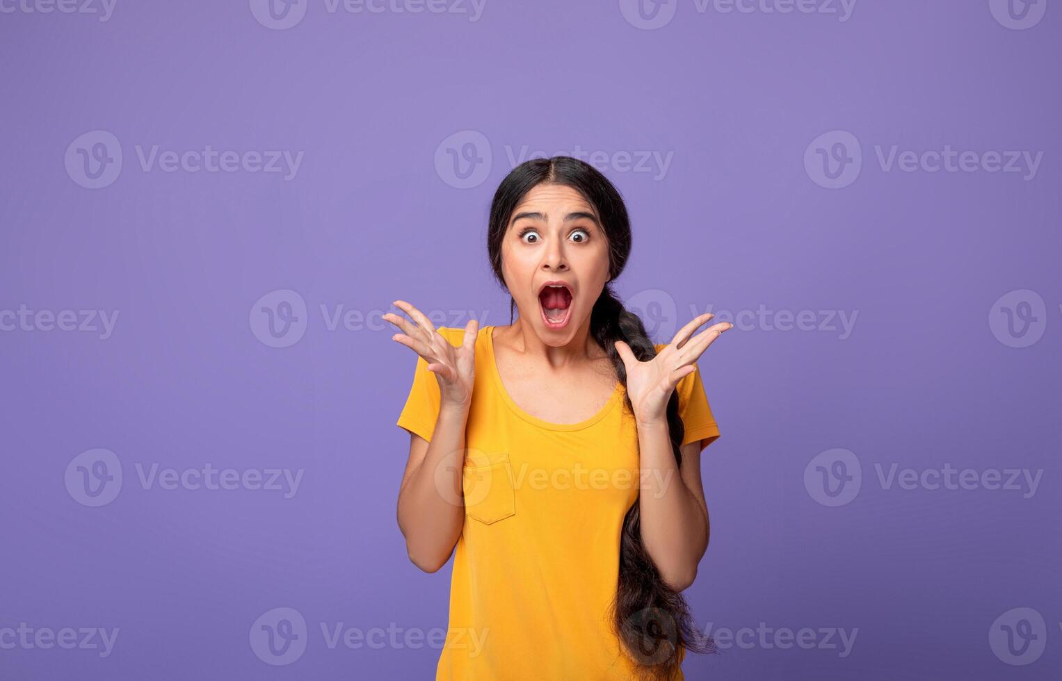 Unbelievable. Portrait of excited overjoyed and shocked young indian woman looking at camera and spreading hands, expressing surprise, screaming with open mouth isolated over purple background photo
